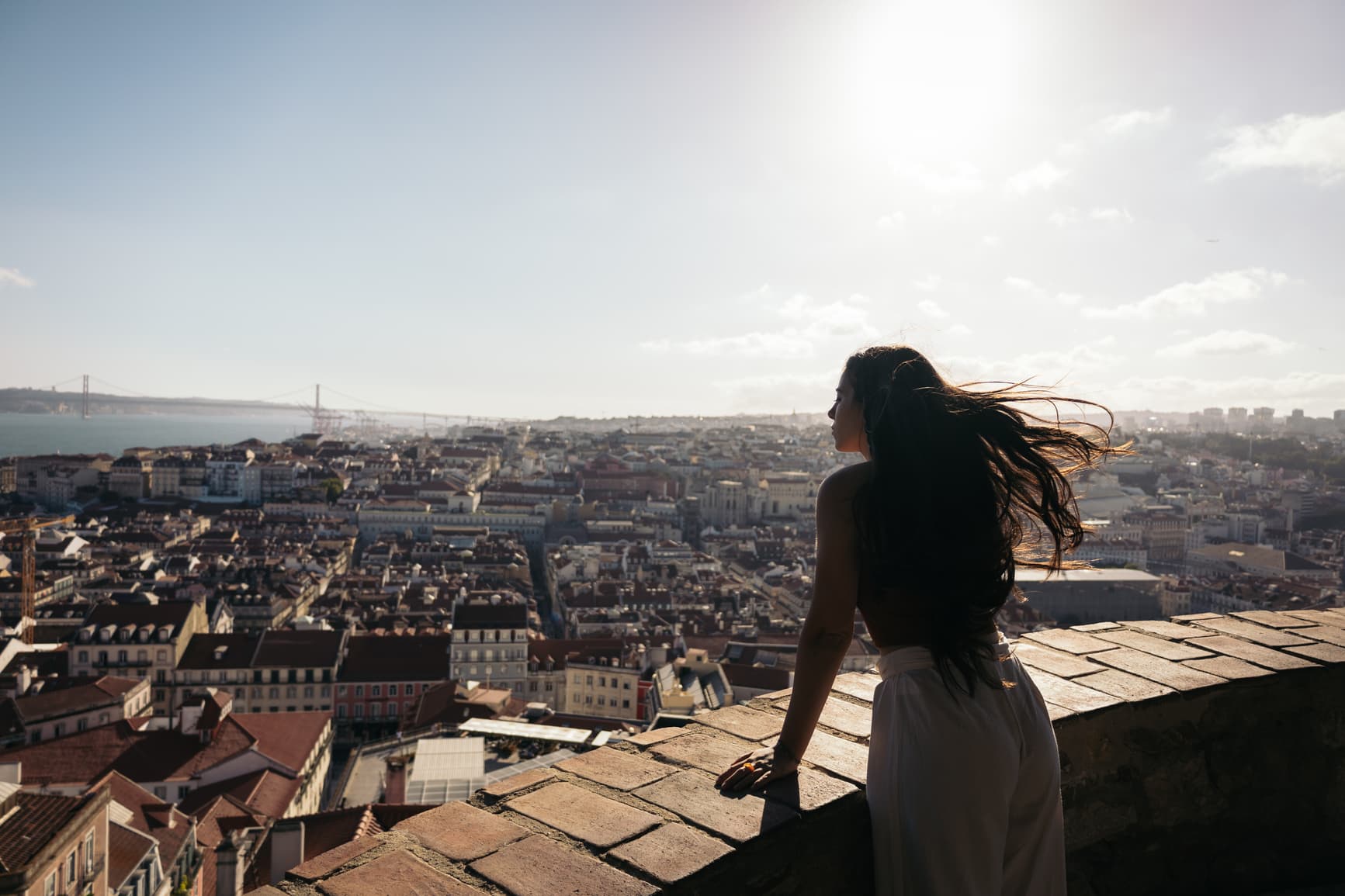 Woman at a viewpoint above Lisbon, hair caught by the wind, looking out over the rooftops toward the river — a moment of unhurried wellbeing